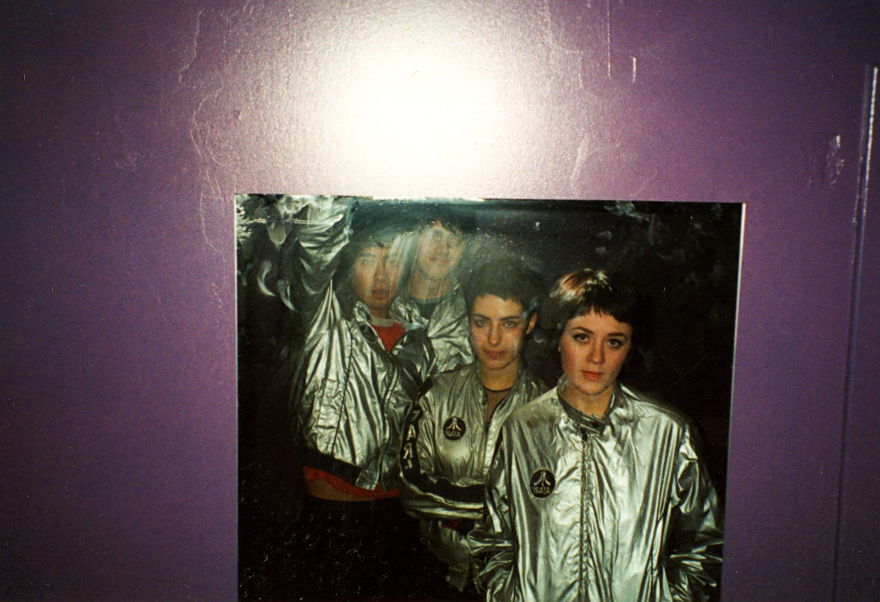 Four people in shiny silver jackets pose together for the photographer, their reflections catching the light against a vibrant purple wall.