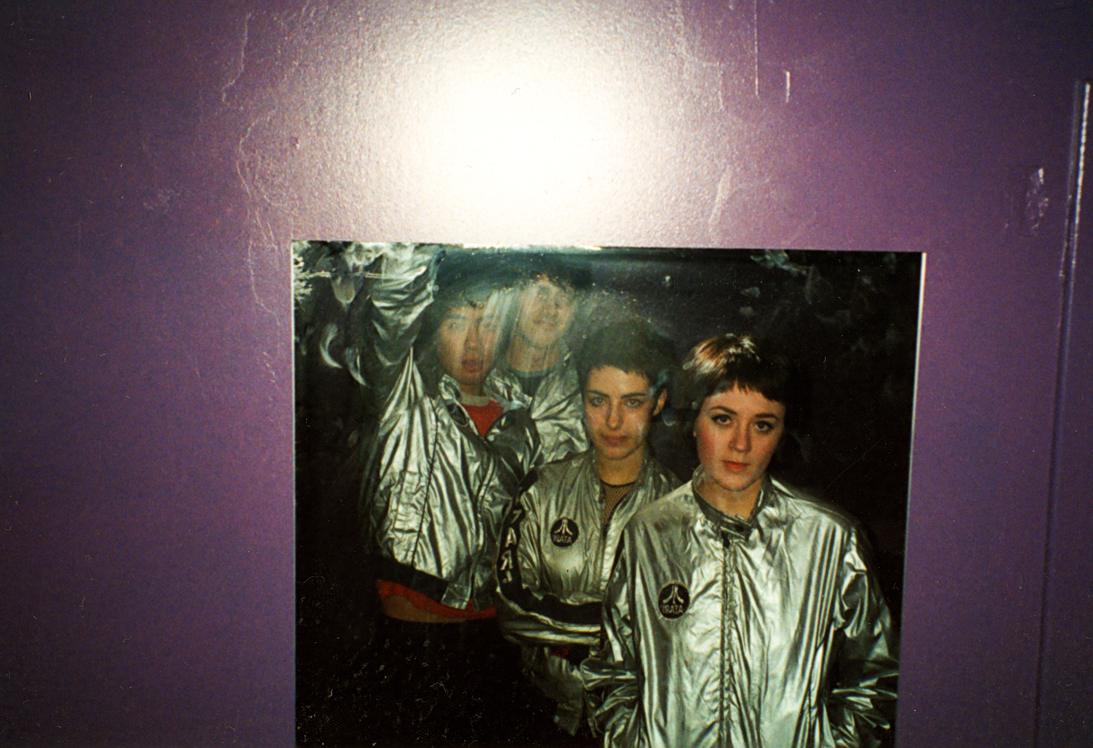 Four people in shiny silver jackets pose together for the photographer, their reflections catching the light against a vibrant purple wall.
