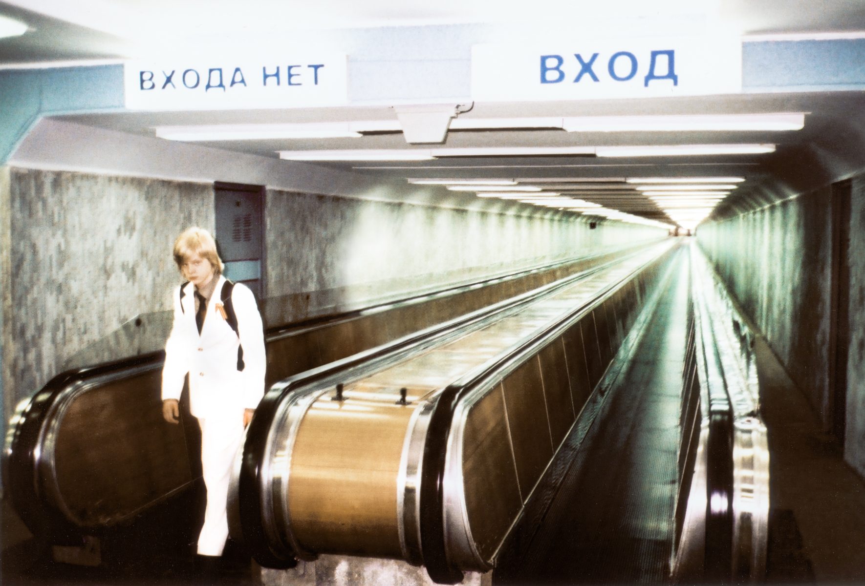 A photographer in white stands on an escalator under Russian signs for No entry and Entrance in a tunnel.