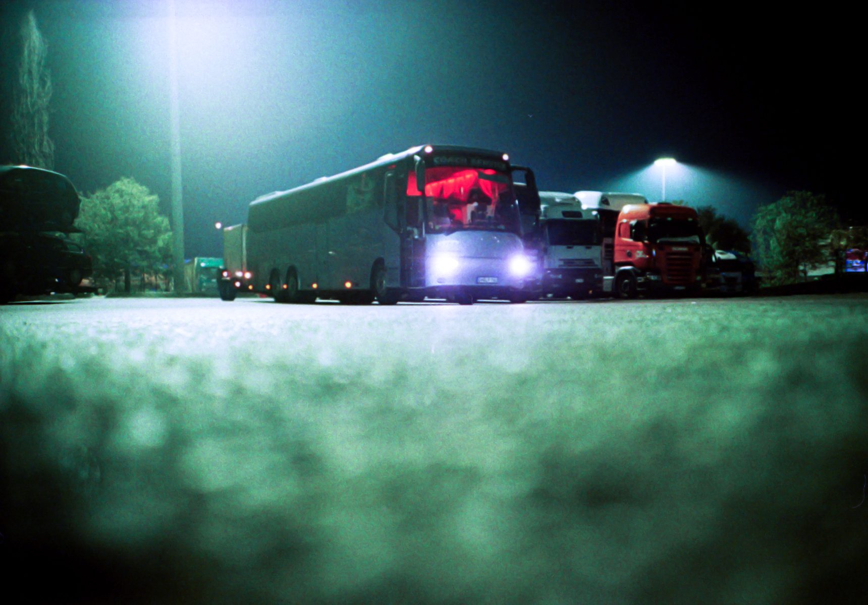 A bus with headlights on is parked in a lot at night, surrounded by trucks under bright streetlights, creating a futuristic scene that would intrigue any photographer.