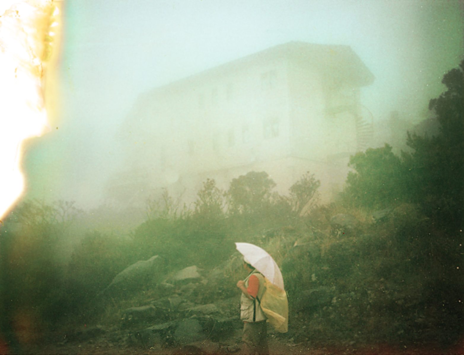 A photographer holding an umbrella stands in a foggy landscape with a blurry building in the background.