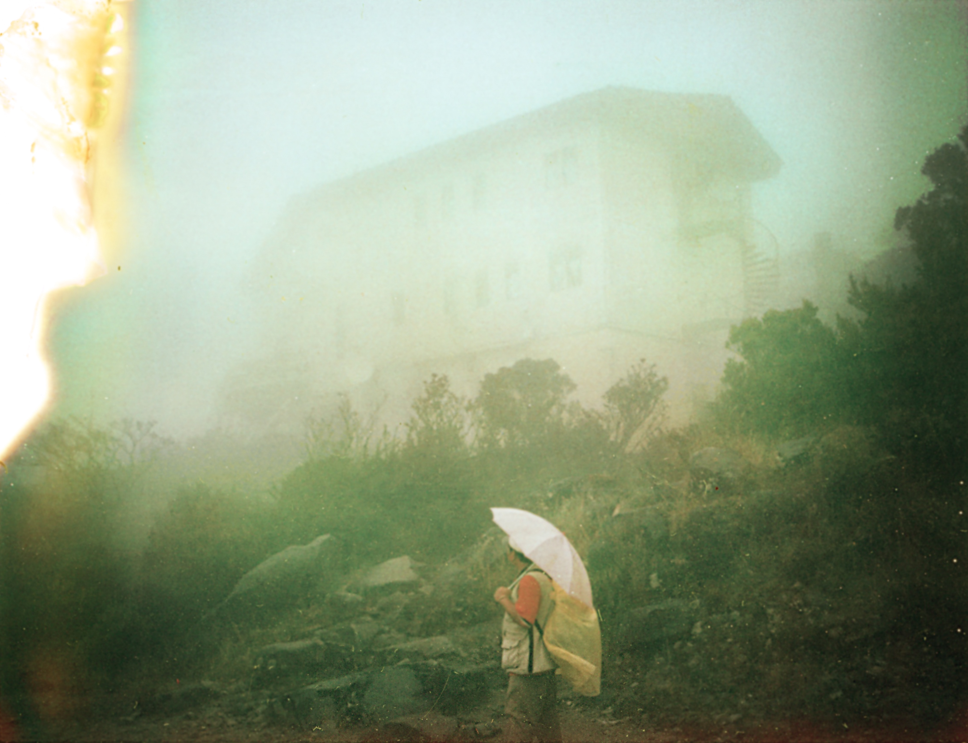 A photographer holding an umbrella stands in a foggy landscape with a blurry building in the background.