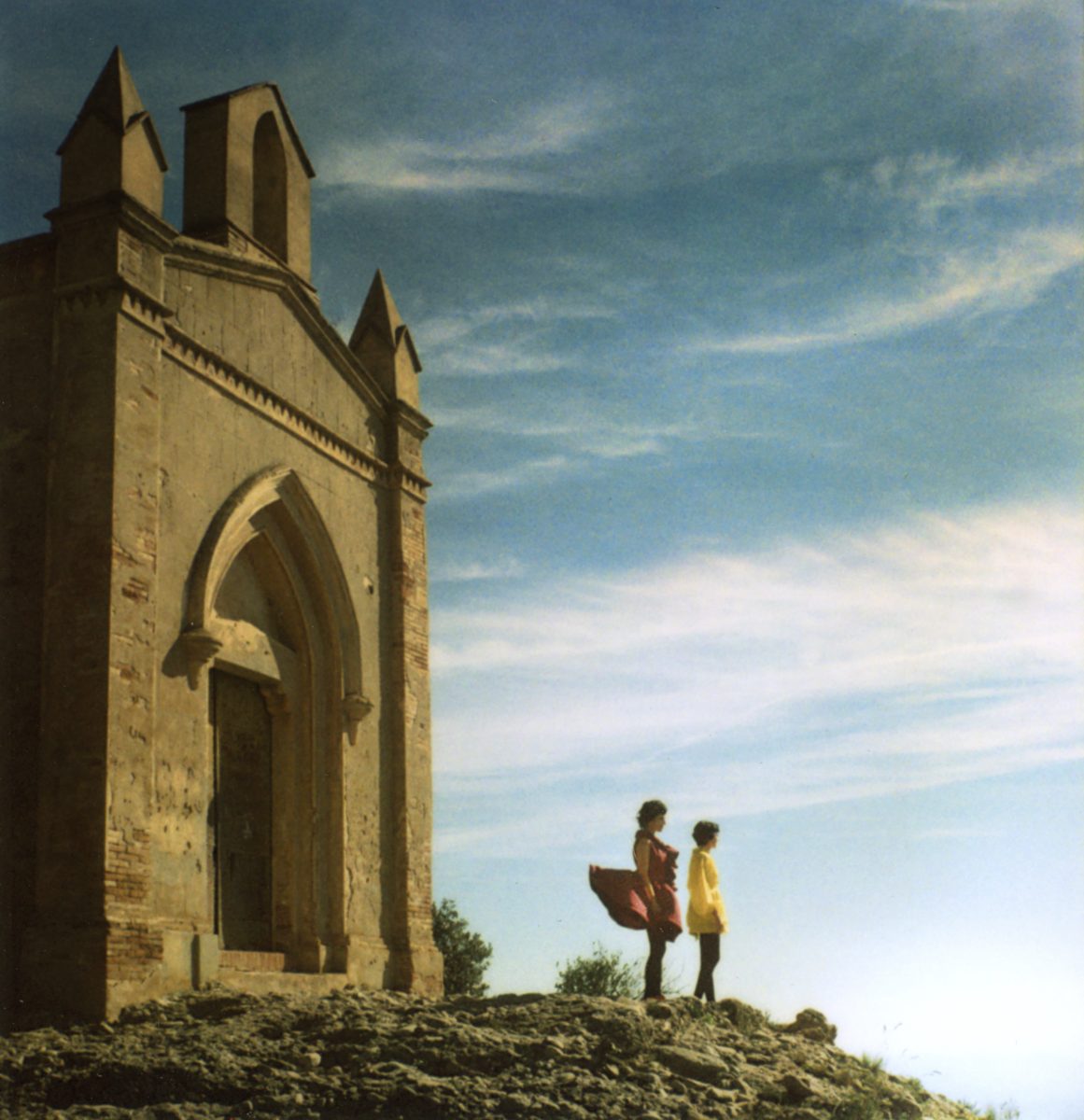 Two people stand near an old stone building on a rocky hill under a blue sky with scattered clouds, as a photographer captures the scene enhanced by drone light painting effects.