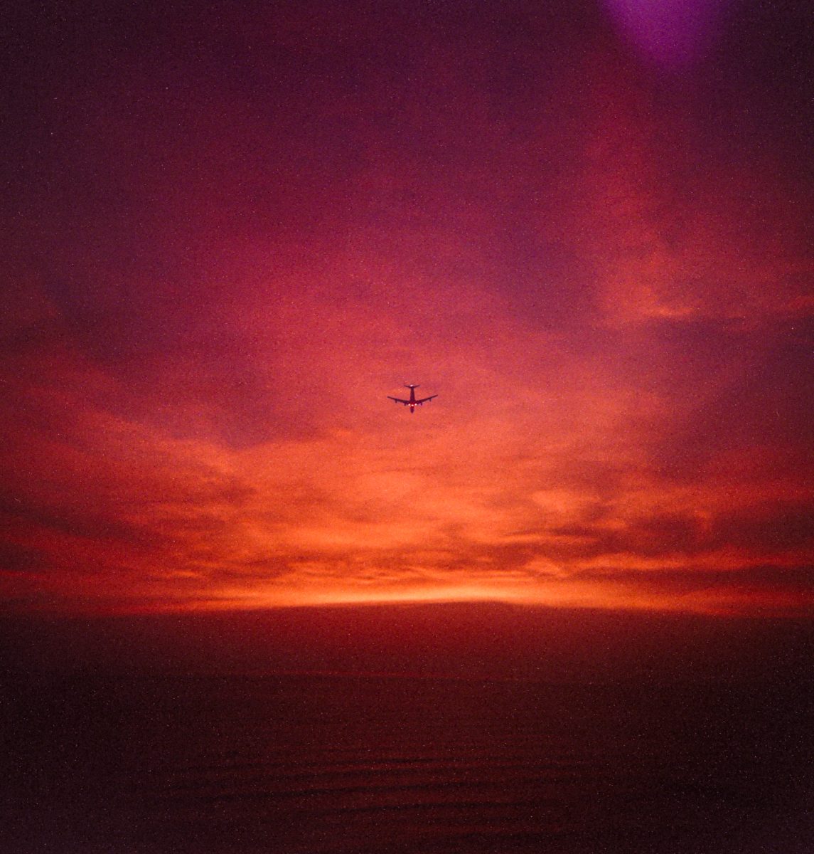 A futuristic airplane soars against a dramatic red and purple sunset sky, capturing a breathtaking scene that any photographer would chase, with hints of drone light painting illuminating the dark horizon.