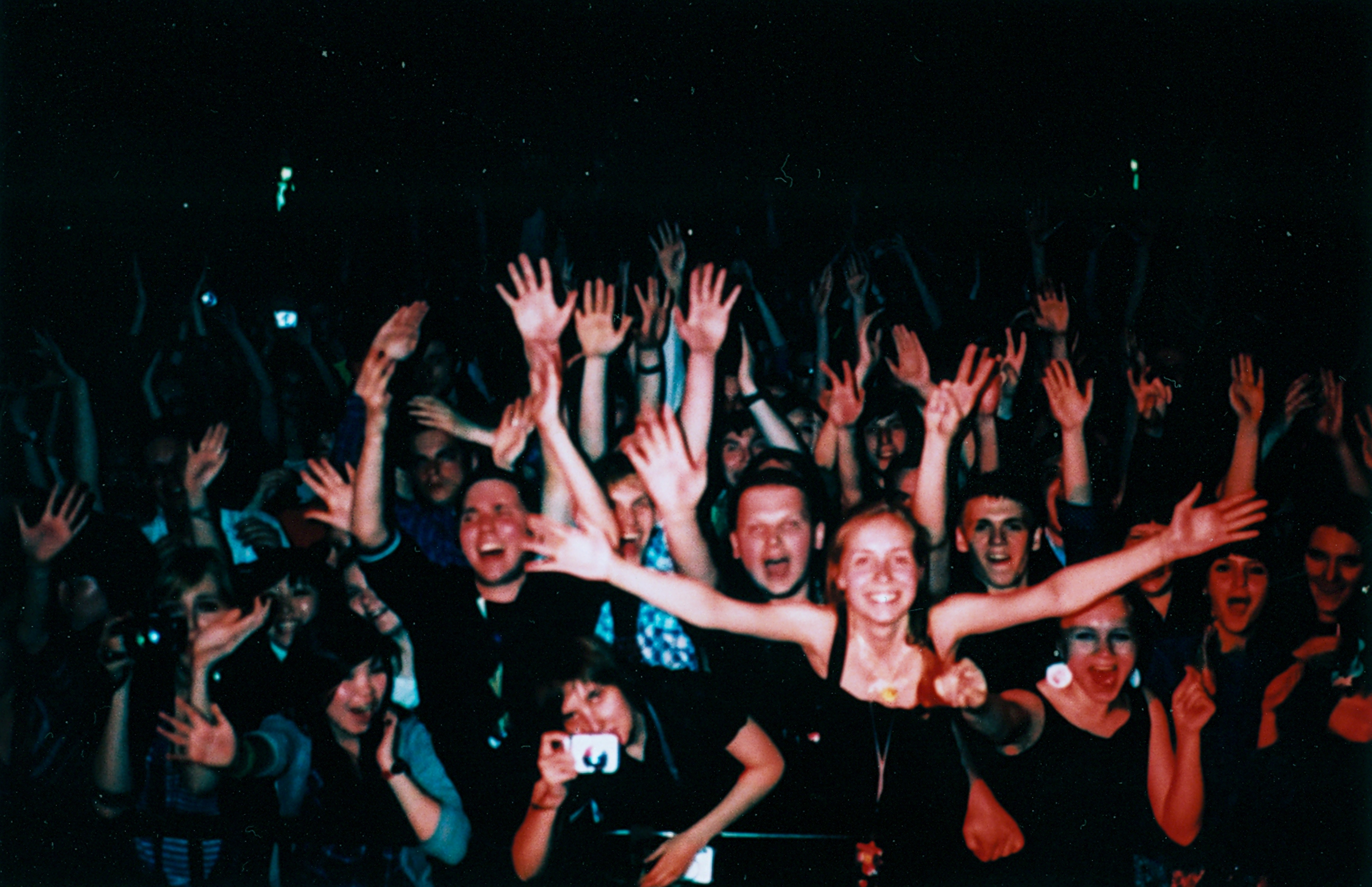 A crowd of people at a concert, smiling and raising their hands toward the camera, as vibrant drone light painting illuminates the scene with artistic flair.