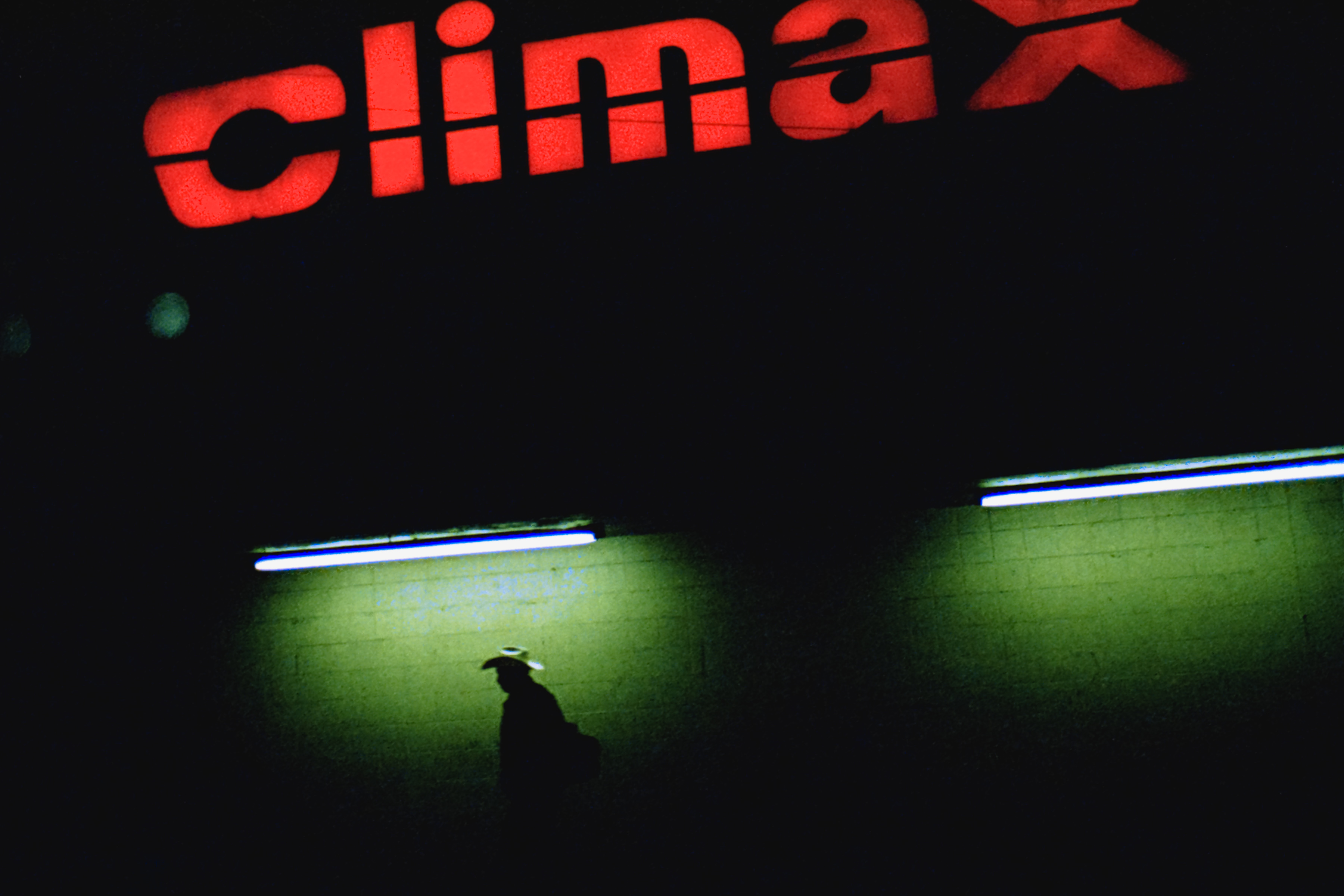 A shadowy figure stands under neon lights below a large, red climax sign at night, creating a futuristic scene that any nighttime landscape photography enthusiast or photographer would admire.