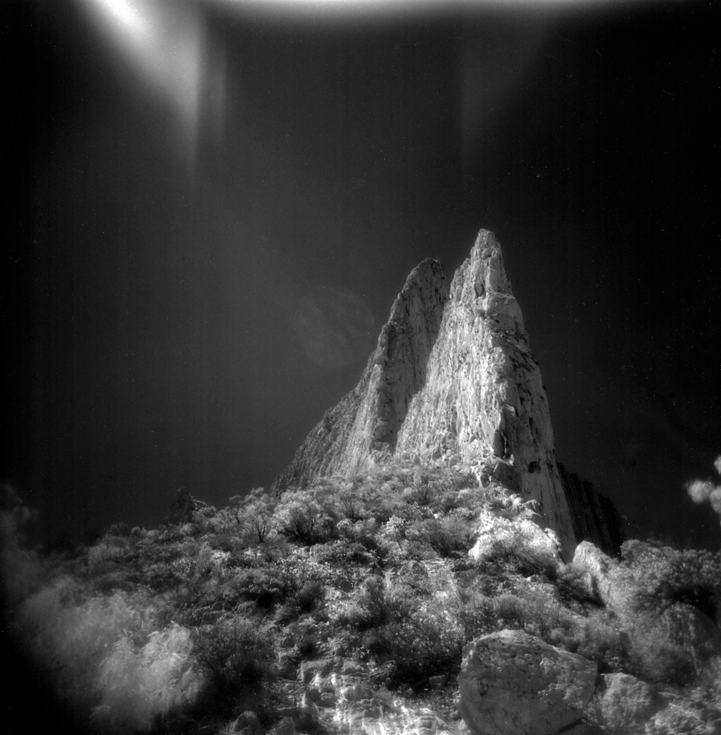 Infrared photo of a steep, rocky mountain peak with bushes under a dramatic, dark sky, evoking a futuristic vibe reminiscent of a fine artist’s drone light painting.