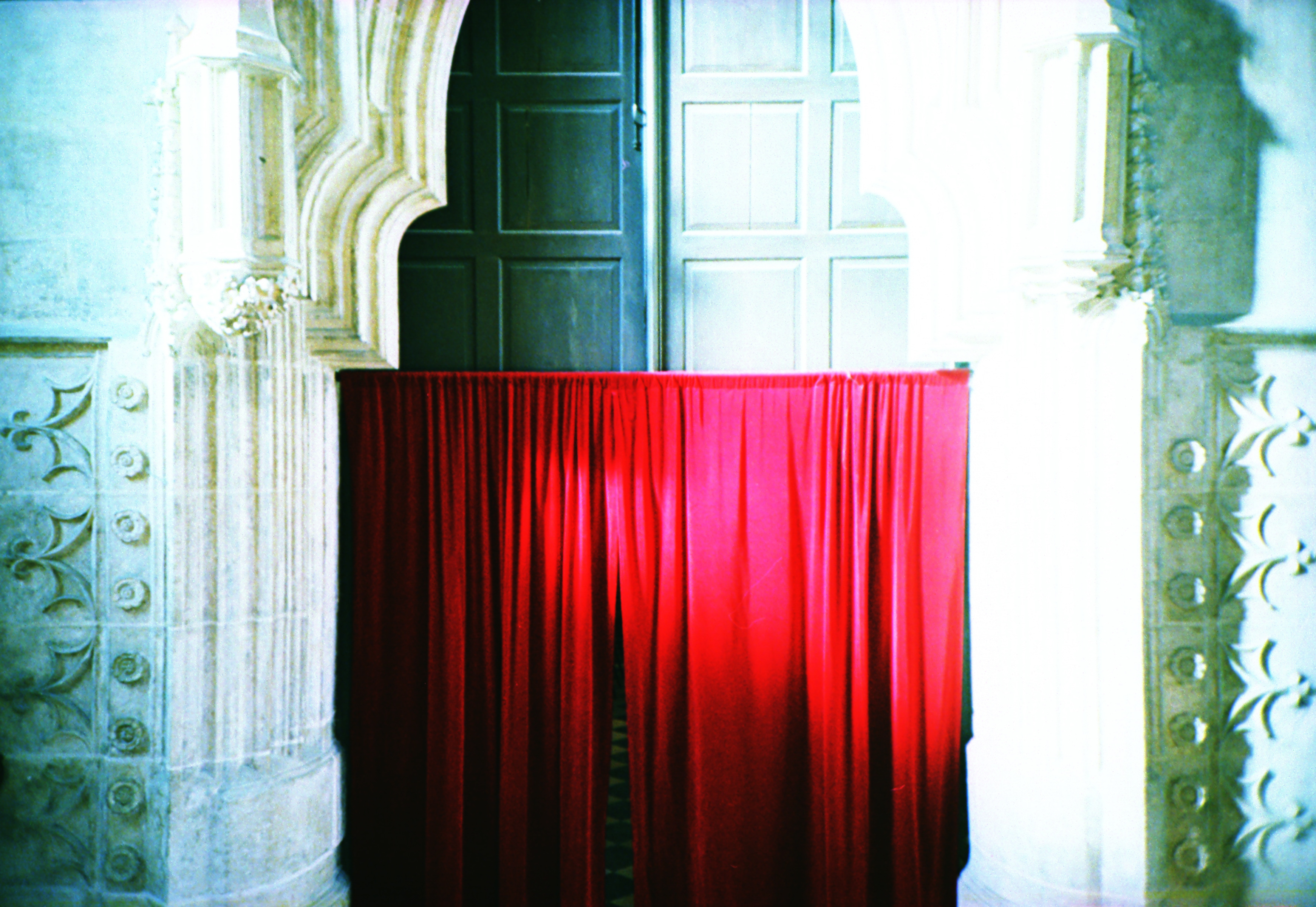A bright red curtain hangs between ornate stone columns in front of a tall, dark wooden door, evoking the dramatic flair of a fine artist’s nighttime landscape photography.