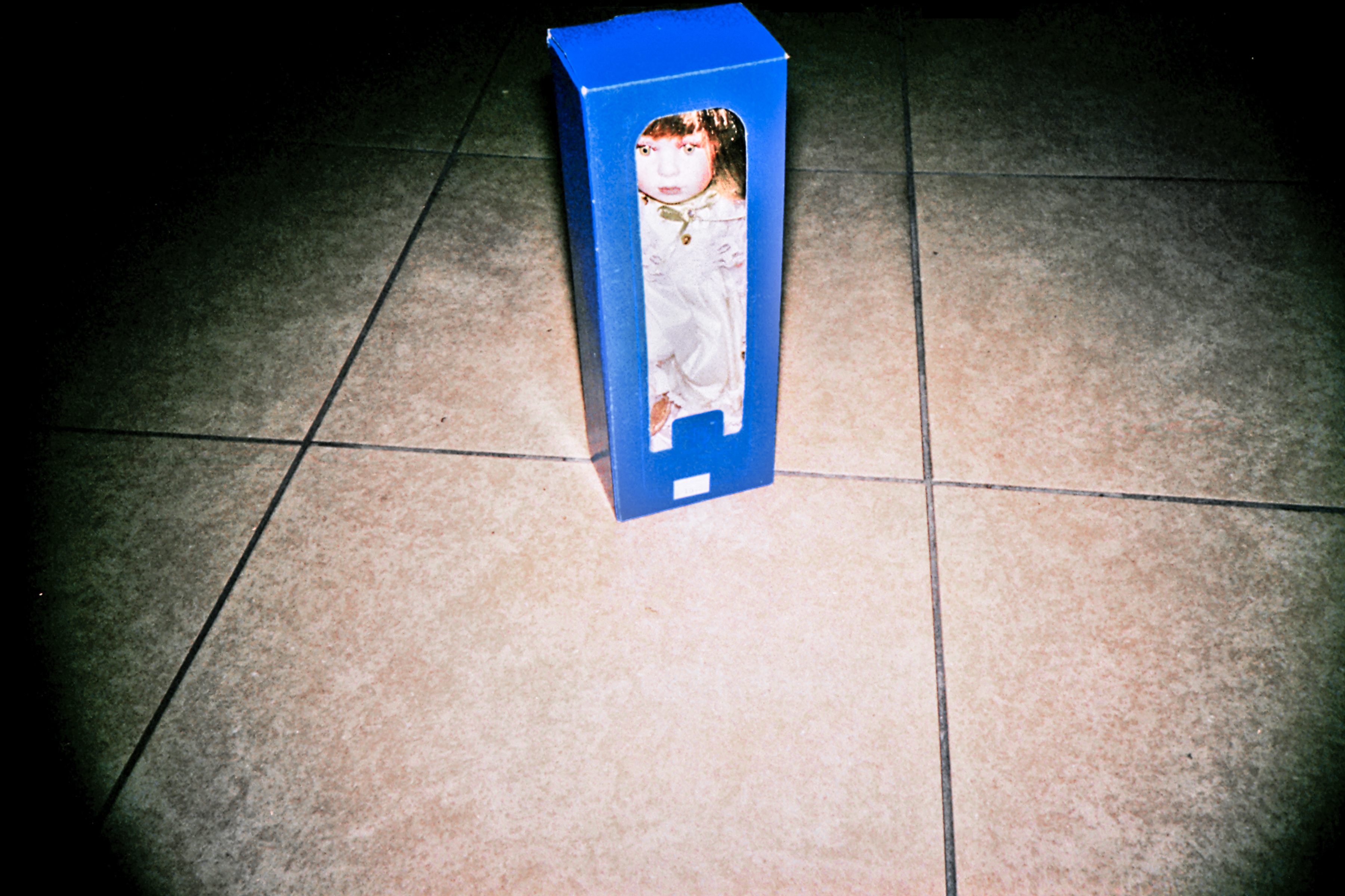A doll in a blue box stands upright on a tiled floor, lit by a camera flash, as if captured by a photographer exploring futuristic scenes.