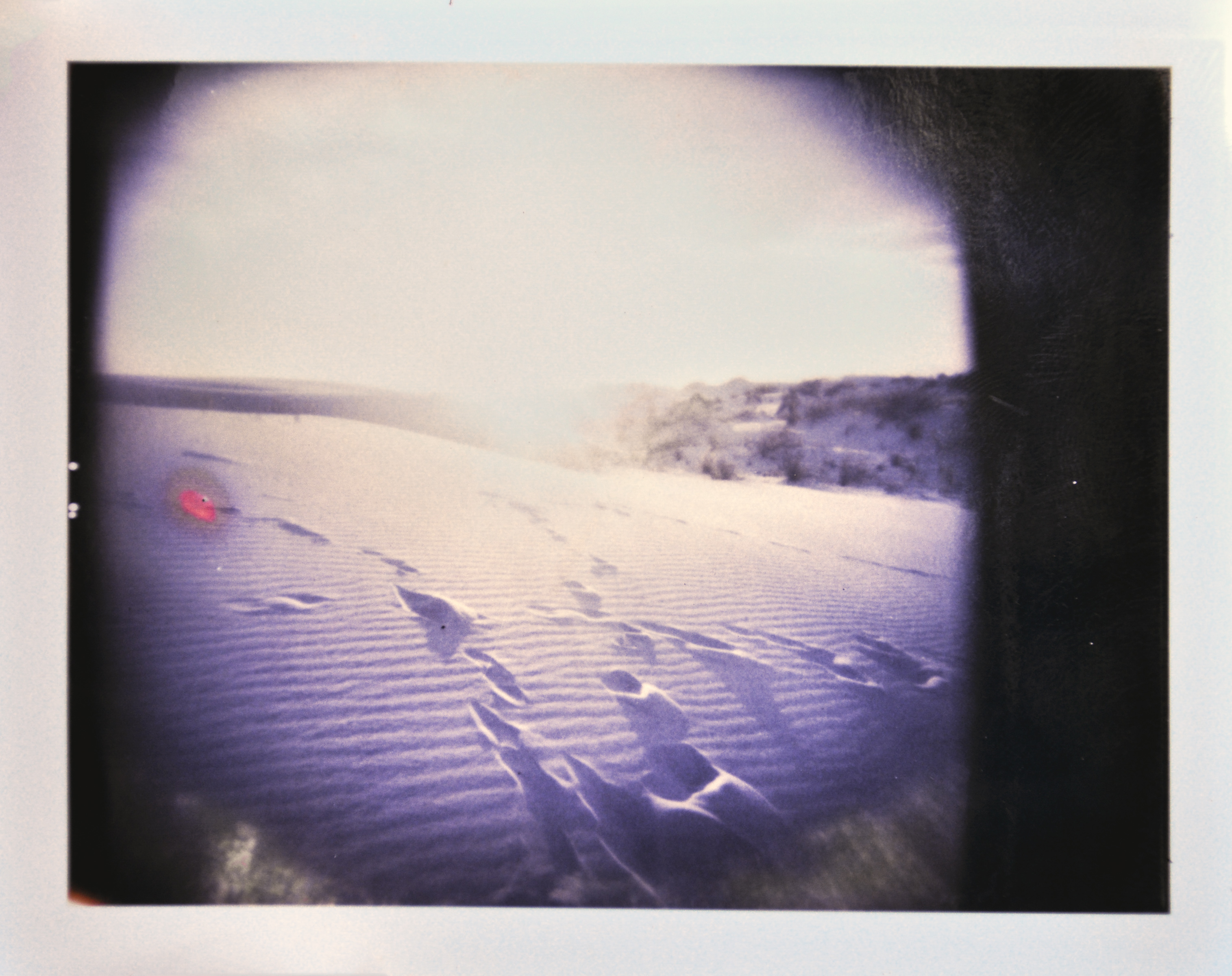 Footprints in rippled sand dunes under a bright sky, with blurred edges and a light leak effect—reminiscent of a fine artist’s nighttime landscape photography.