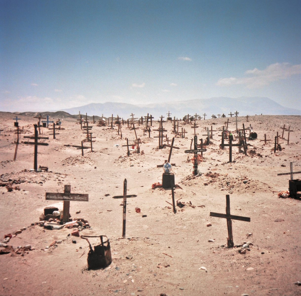 A barren desert cemetery with many wooden crosses marking graves under a clear sky, illuminated by ethereal patterns of drone light art.
