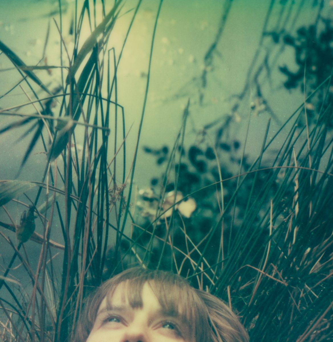A person’s face peeking through tall grass by a reflective, green-tinted water surface in a nighttime landscape photography scene.