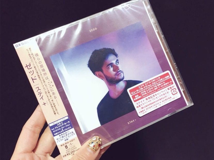 Hand holding a Zedd Stay + CD with Japanese text and a purple-toned, futuristic album cover reminiscent of nighttime landscape photography.