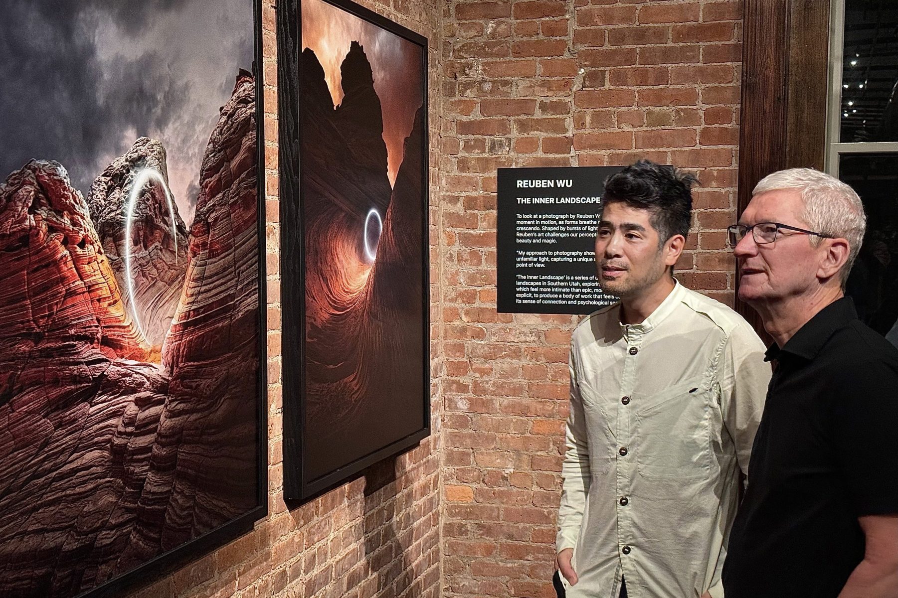 Two people view dramatic landscape photography by a renowned photographer in a brick-walled gallery.