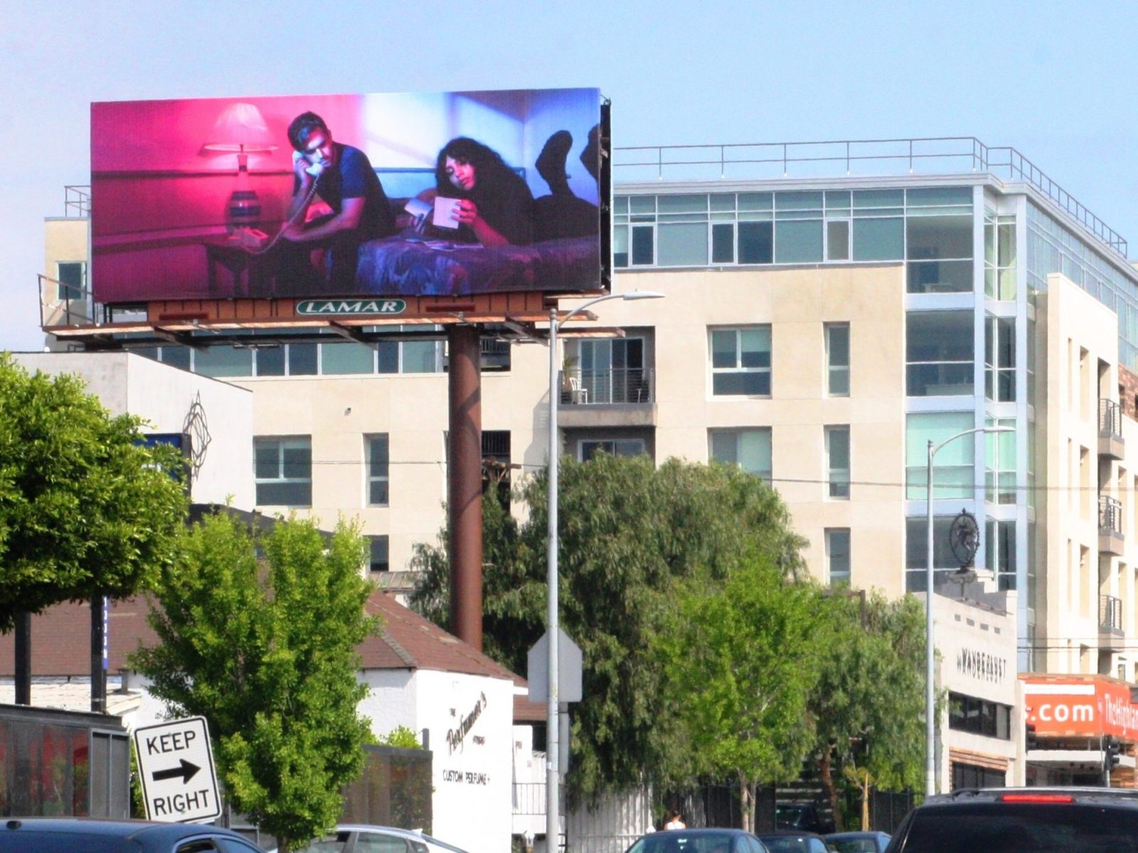 Billboard shows two people relaxing on a couch with warm lighting, above a city street with buildings and trees, capturing a touch of futuristic charm reminiscent of nighttime landscape photography.