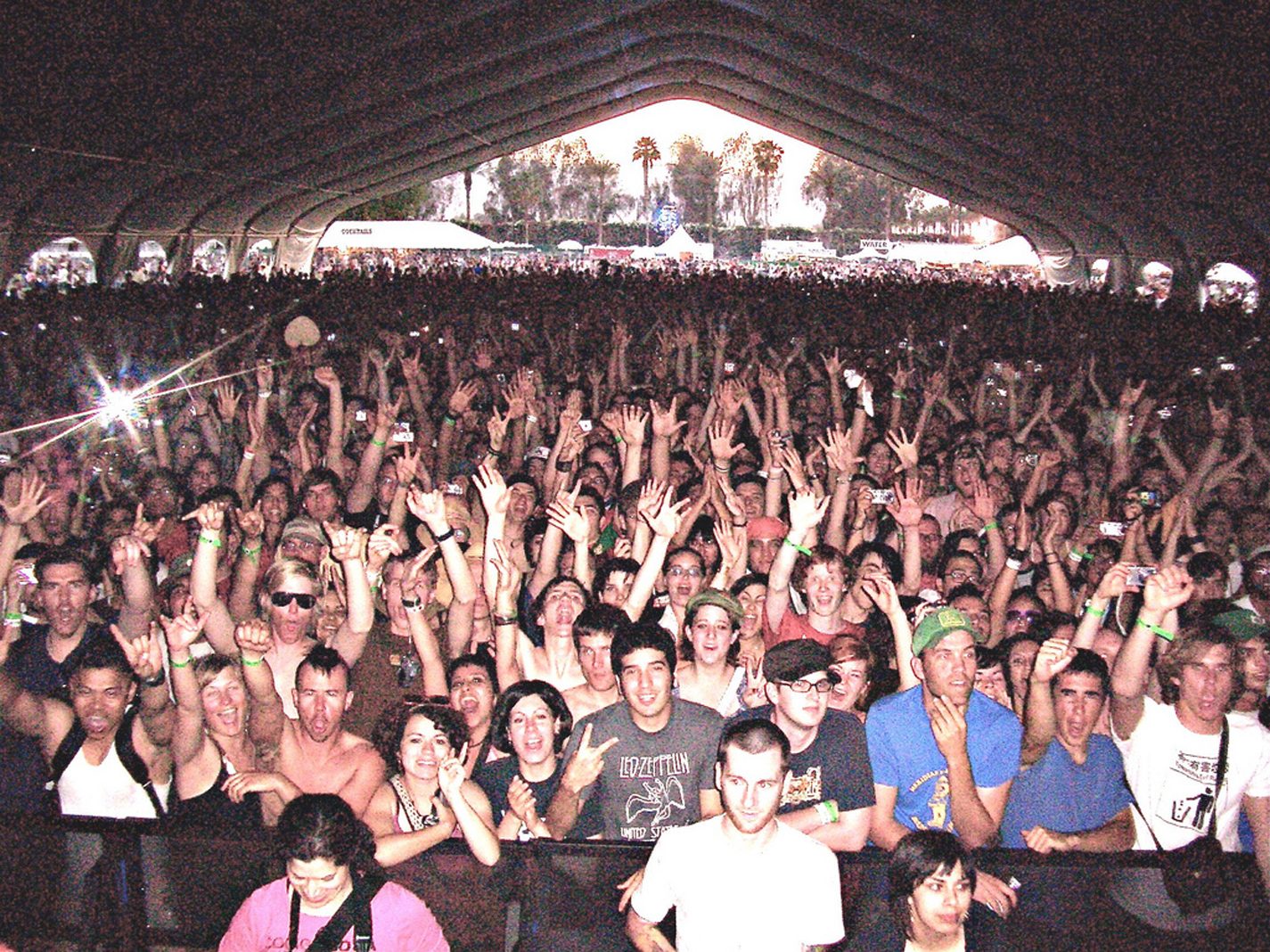 A large crowd at a concert, many people with hands raised, fills a futuristic tent with vibrant lights in the background.