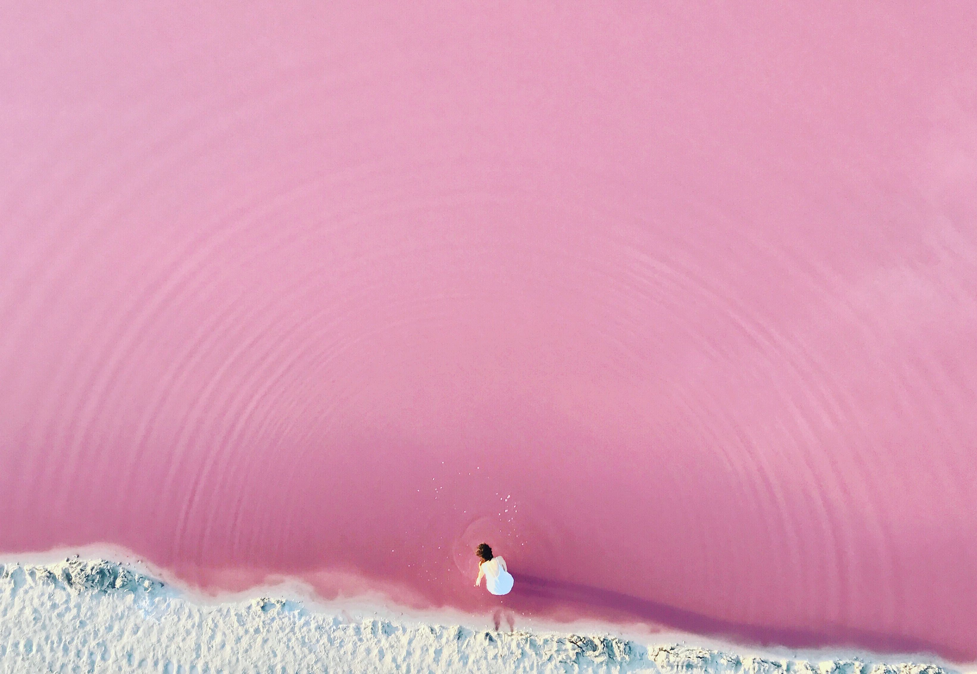 A fine artist in white stands at the edge of a pink lake with ripples, seen from above.