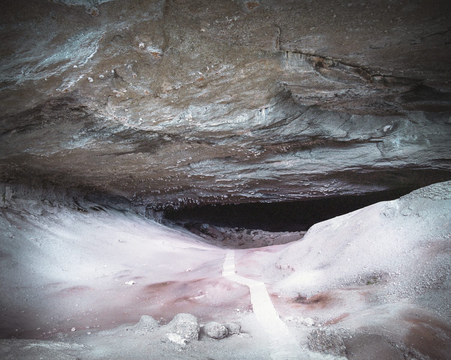 A narrow path leads into the dark entrance of a large, rocky cave with a low ceiling, perfect for a fine artist or photographer seeking dramatic inspiration.