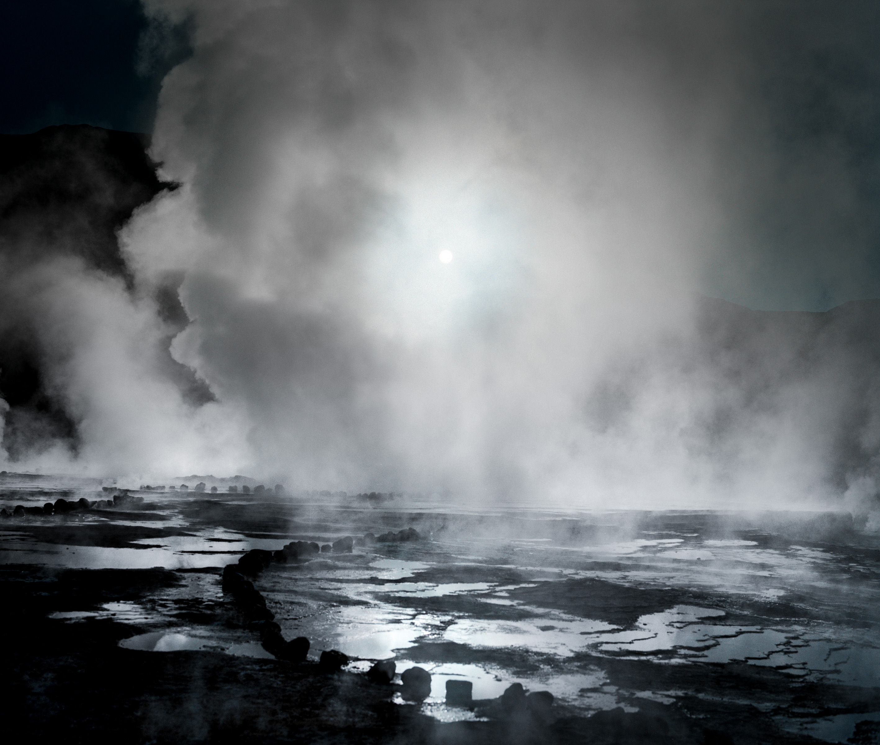Thick steam rises from a geothermal hot spring, partially obscuring the sun and rocky landscape, creating a mystical scene perfect for a photographer or fine artist to capture in striking detail.