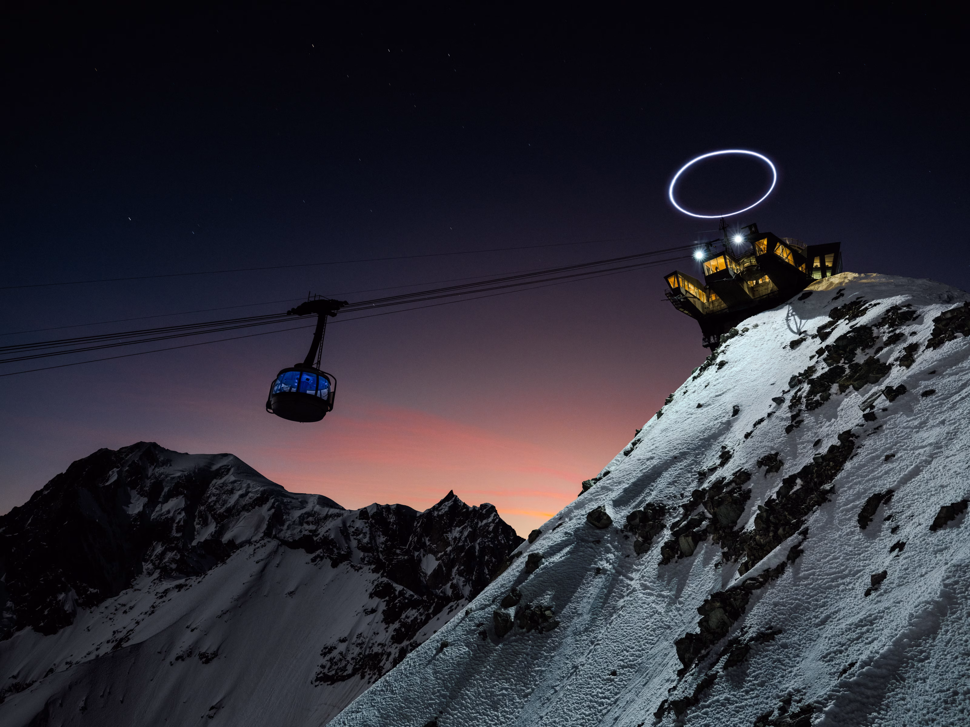 A cable car ascends snowy Monte Bianco at dusk on the Skyway, passing a lit station crowned with a circular light above it.