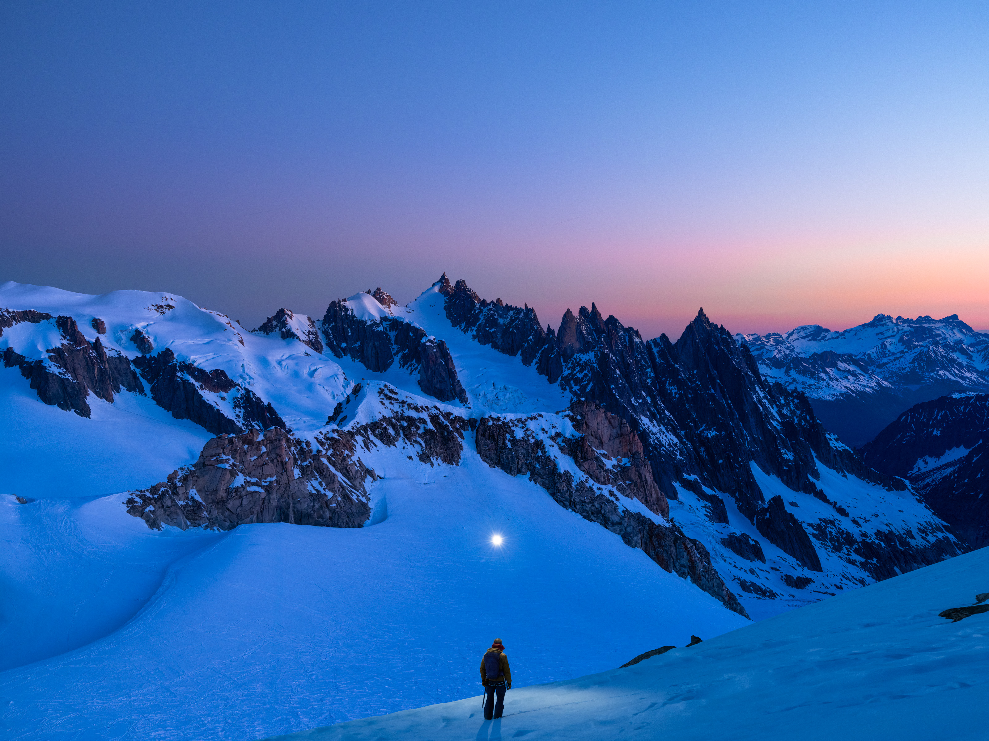 A person stands on a snowy mountain slope at dusk, overlooking jagged peaks under a colorful sky—perfect for nighttime landscape photography.