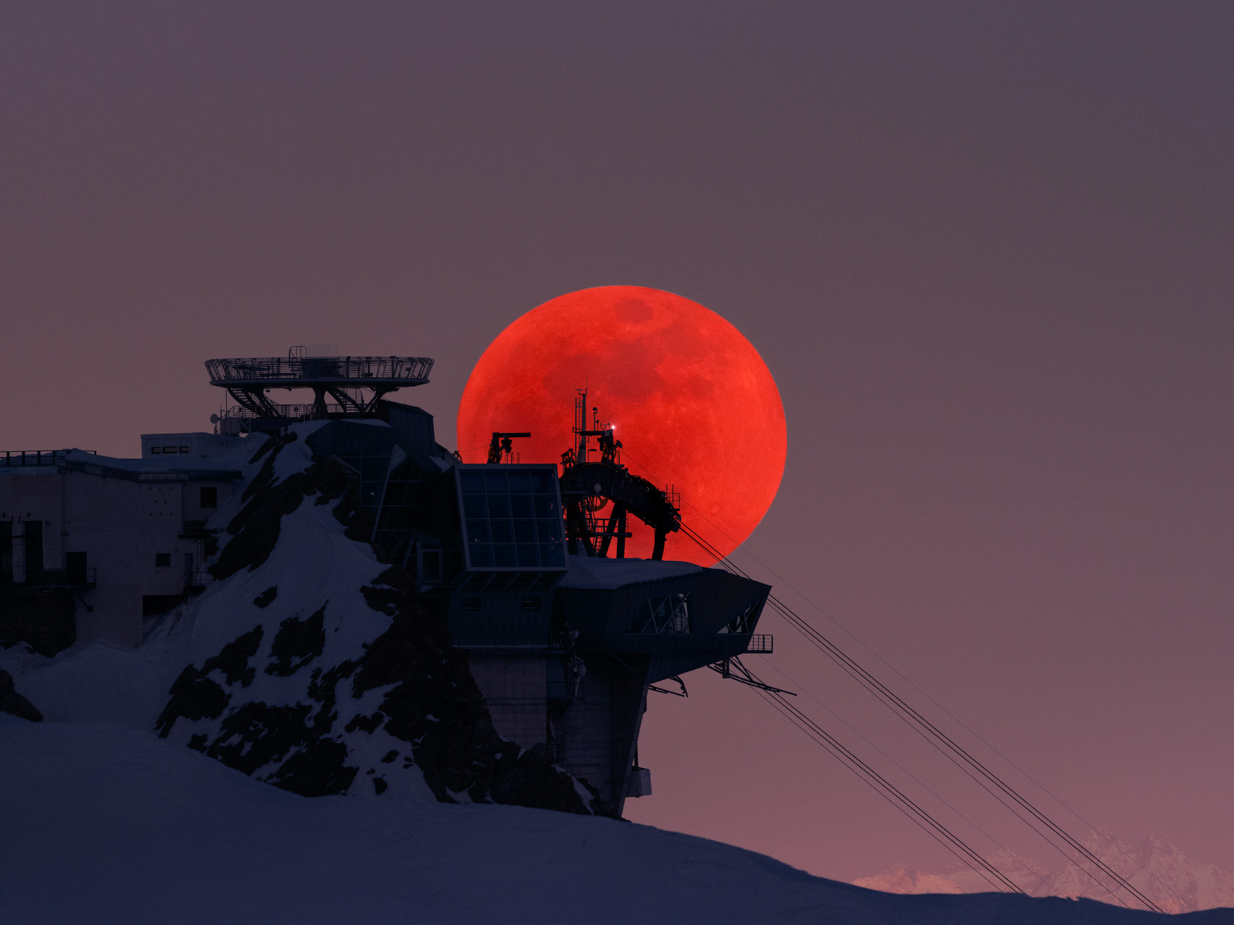 A large red moon rises behind a snowy mountain building and ski lift at dusk, captured in stunning nighttime landscape photography.