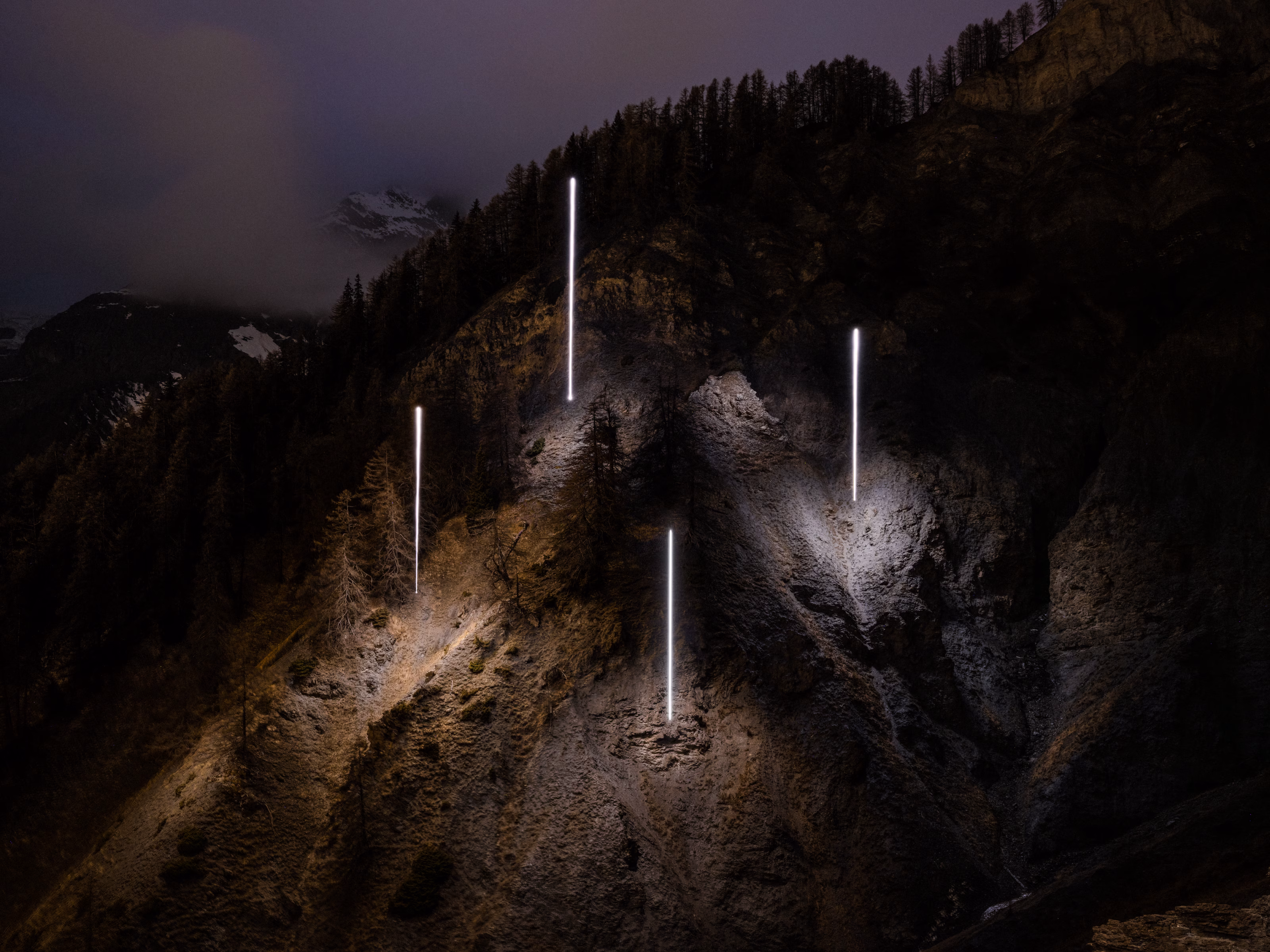 Vertical light beams illuminate a rocky, forested mountain slope at night under a cloudy sky, with the Skyway Monte Bianco cable car visible in the distance.